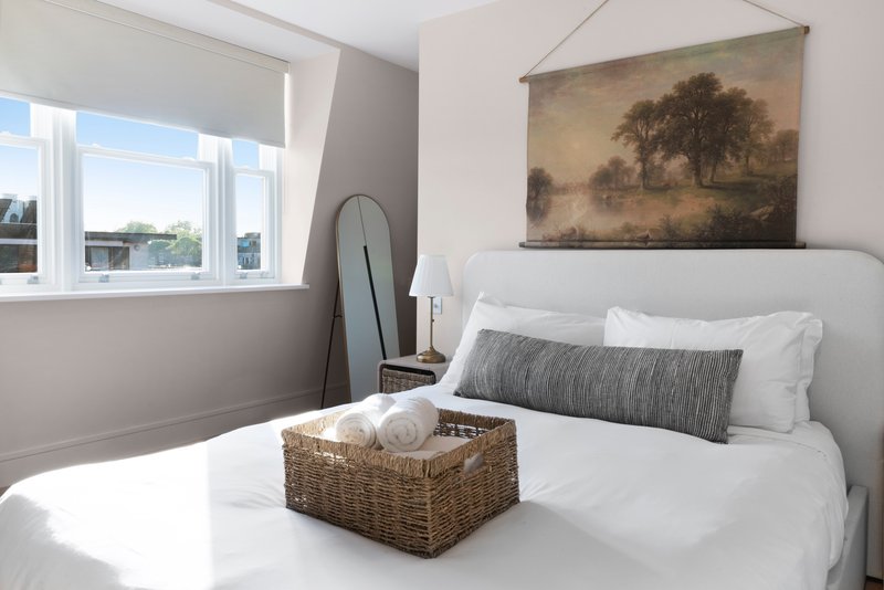 Bright bedroom with white bed, grey cushion, landscape artwork, and window with city view