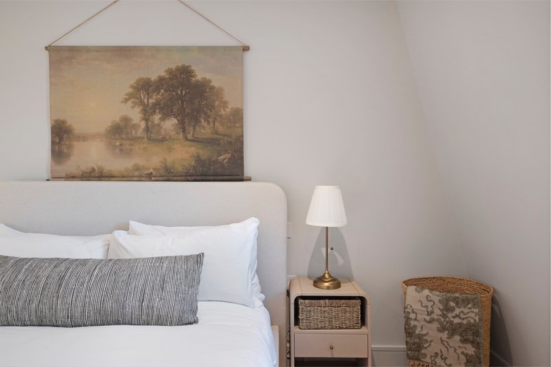 Bedroom with white upholstered headboard, white bedding, grey cushion, framed landscape artwork above bed, and bedside lamp