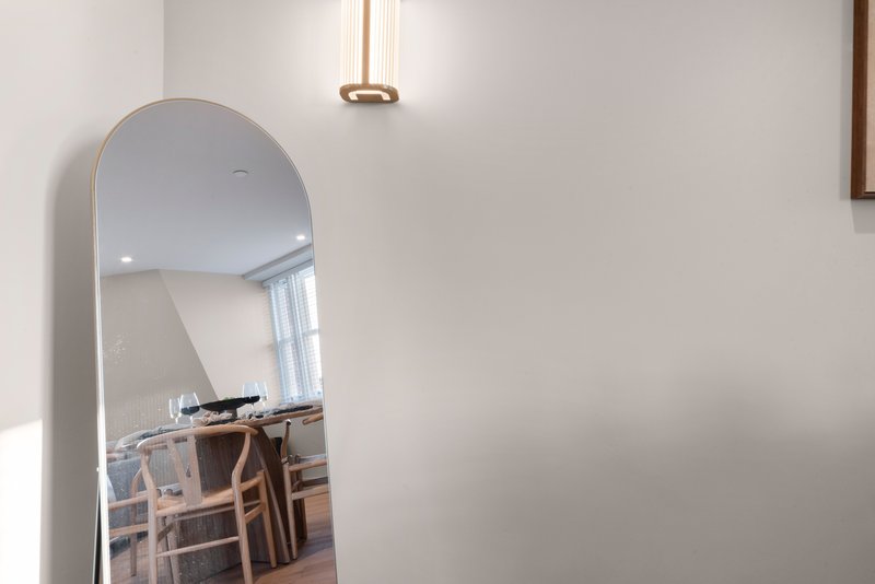 Arched mirror leaning against white wall, reflecting dining area with wooden chairs
