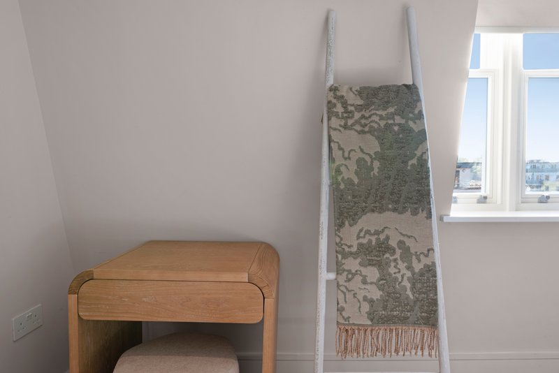 Bedroom corner with wooden desk, decorative tapestry on ladder, and window view