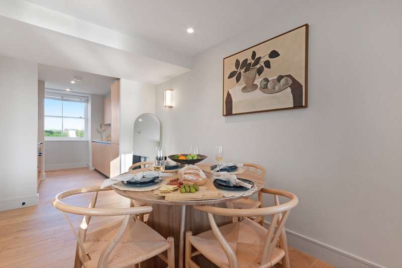 Bright dining room with round wooden table, cross-back chairs, place settings, and kitchen visible beyond