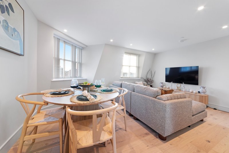 Open-plan living and dining area with wooden dining table, gray sofa, TV, and natural light from windows