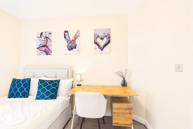 Bedroom with double bed, blue patterned pillows, wooden desk, white chair and wall art