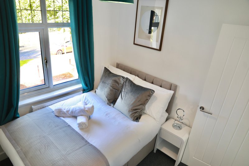 Bright bedroom with double bed, white bedding, grey and white pillows, teal curtains and small bedside table