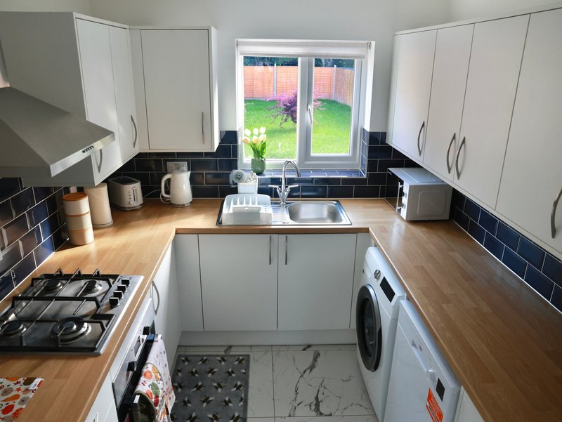 Modern kitchen with white cabinetry, black tile splashback, wooden worktops, appliances and garden view through doors