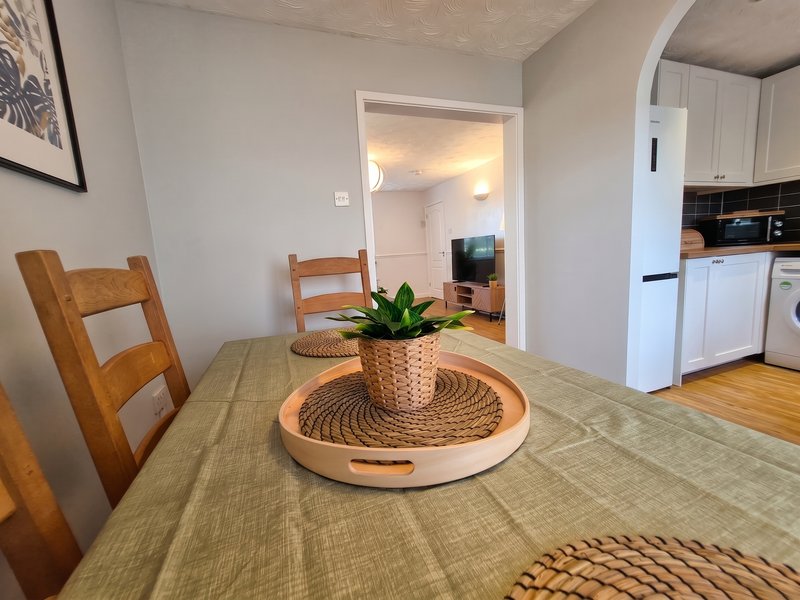 Open-plan dining and kitchen area with wooden chairs, dining table, potted plant, and white appliances visible