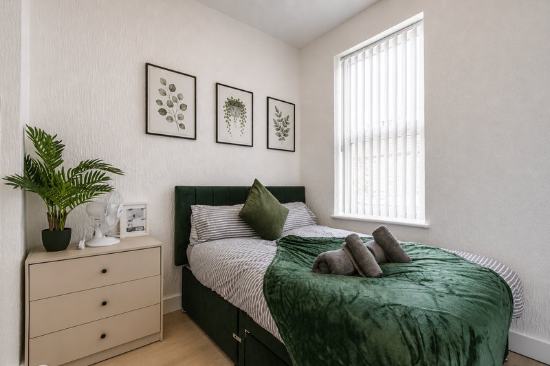 Modern bedroom with dark green bed, cream dresser, potted plant, and framed botanical wall art