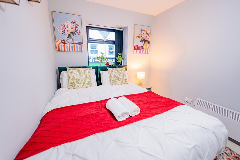 Bright bedroom with double bed, red runner, patterned cushions and framed floral artwork