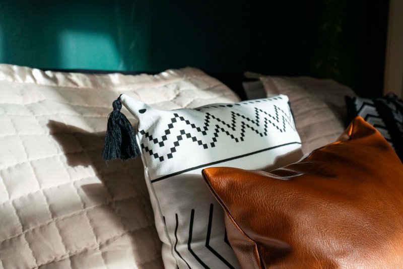 Bed with white quilted duvet, patterned cushions with tassels and geometric designs, cognac leather bag
