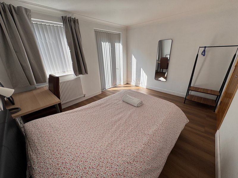 Spacious bedroom with double bed, wooden flooring, desk, mirrors and vertical blinds