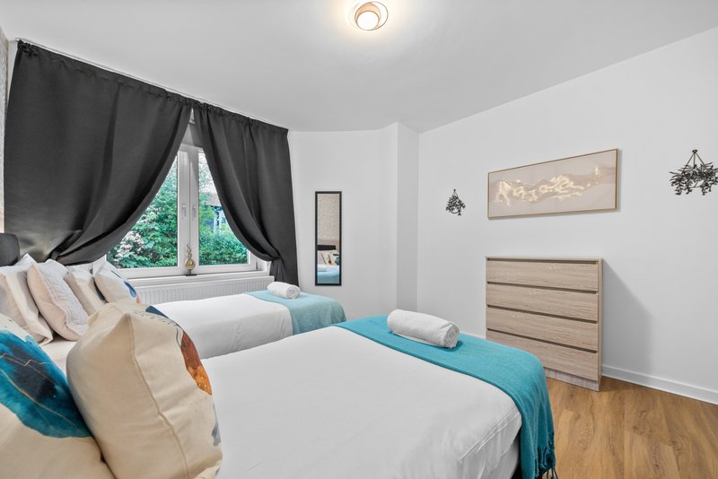 Bright bedroom with two beds, blue bedding, black curtains and wooden flooring
