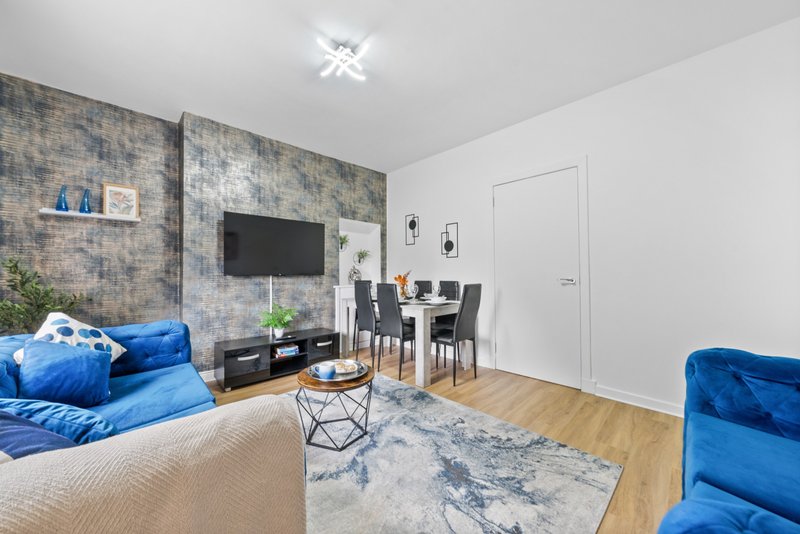 Modern open-plan living room with blue sofa, dining table, TV wall, and wooden flooring