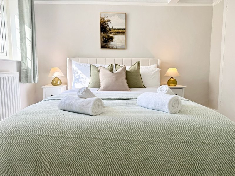 Modern bedroom with neatly made bed, green throw, decorative pillows, rolled towels, and bedside lamps