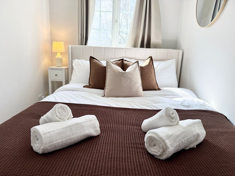 Bedroom with double bed, brown bedspread, rolled towels, decorative pillows and bedside lamp