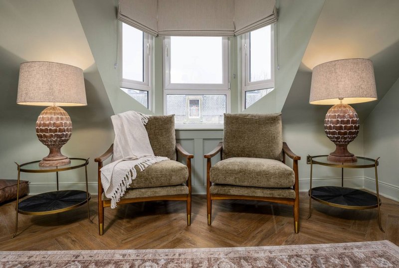 Bright sitting room with two upholstered chairs, table lamps, and bay windows overlooking street