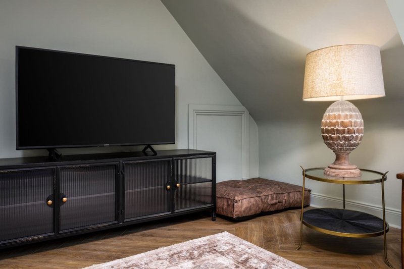 Attic bedroom with TV, black media console, patterned rug, ottoman, and table lamp
