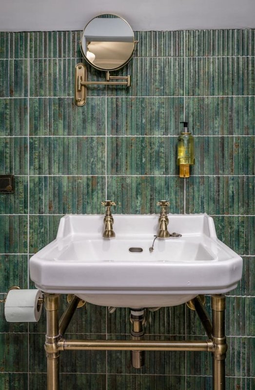 Bathroom with white pedestal sink, brass fittings, round mirror and green tile wall