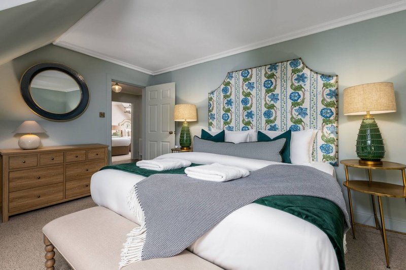 Bedroom with queen bed, blue patterned headboard, wooden dresser, round mirror and table lamps
