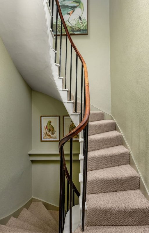 Interior staircase with wooden handrail, black metal balusters, and beige carpeted steps