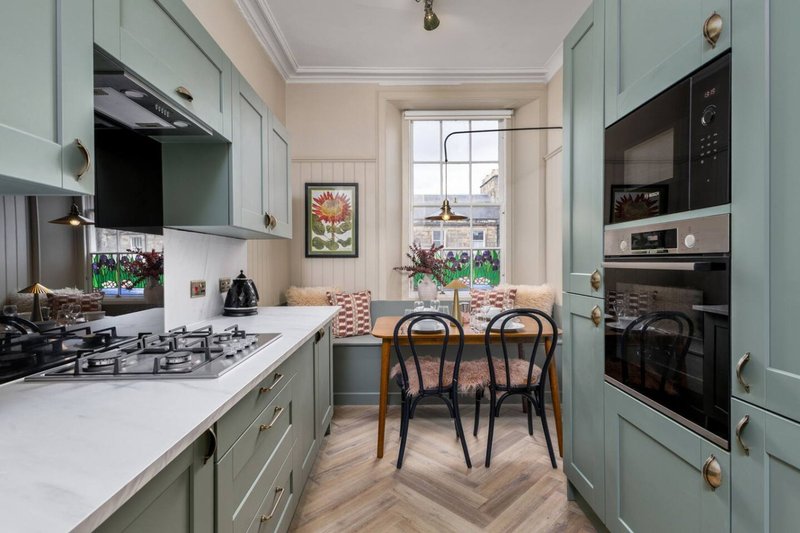 Modern galley kitchen with sage green cabinetry, white counters, dining table and chairs by window