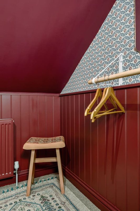 Small burgundy dressing room with wooden stool, gold hangers and patterned wallpaper