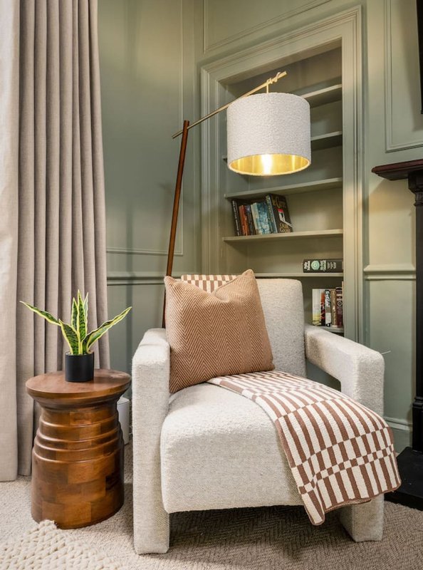 Cream upholstered armchair with cushion and throw blanket, wooden side table with plant, floor lamp, and built-in shelving behind