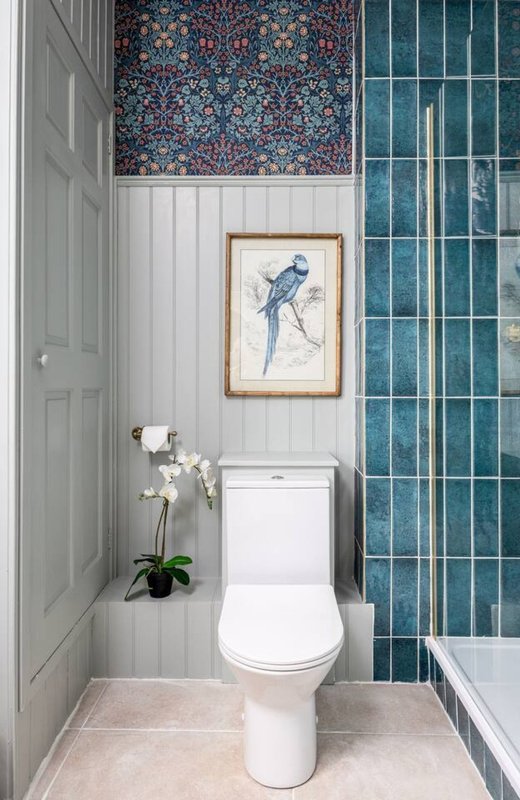 Compact bathroom with white toilet, teal tile shower enclosure, framed bird artwork, and potted orchid