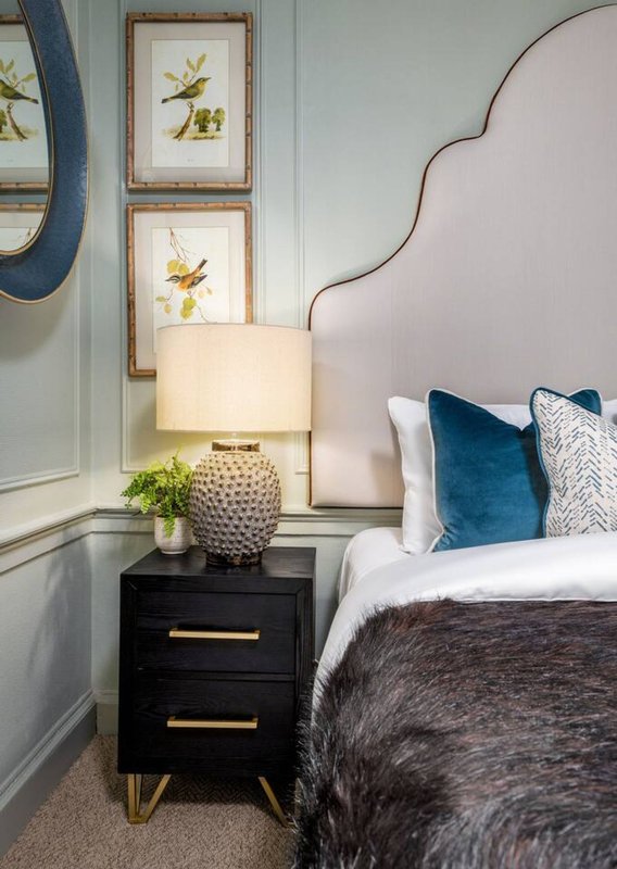 Bedroom corner with upholstered headboard, black nightstand, lamp, and framed botanical wall art