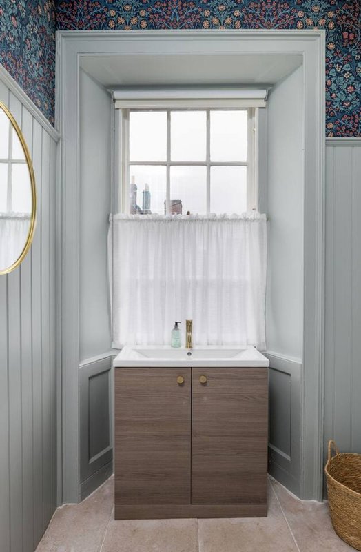 Bathroom with wooden vanity, white curtained window, patterned wallpaper and paneling
