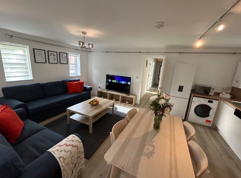 Open-plan living room with dark blue sofa, wooden coffee table, TV, and kitchen area with white appliances