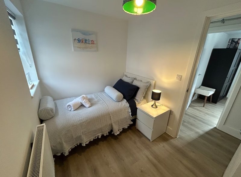 Small bedroom with single bed, white bedding, black cushions, wooden floor and bedside lamp