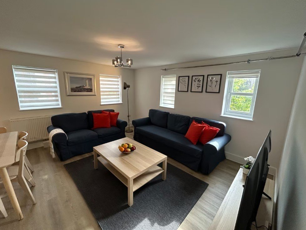 Spacious living room with black corner sofa, light wood coffee table, red cushions and multiple windows
