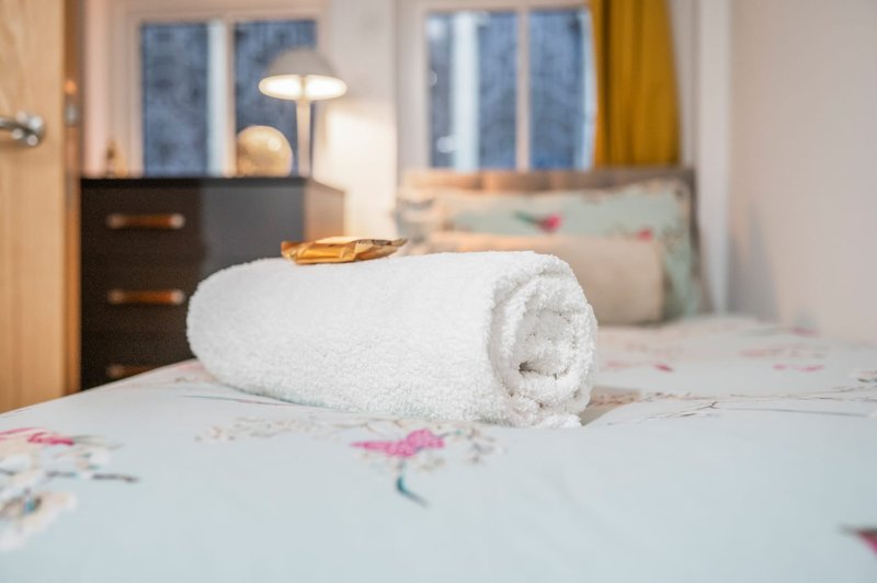 Bedroom with rolled white towel on bed, dark dresser, lamp, and windows with yellow curtains