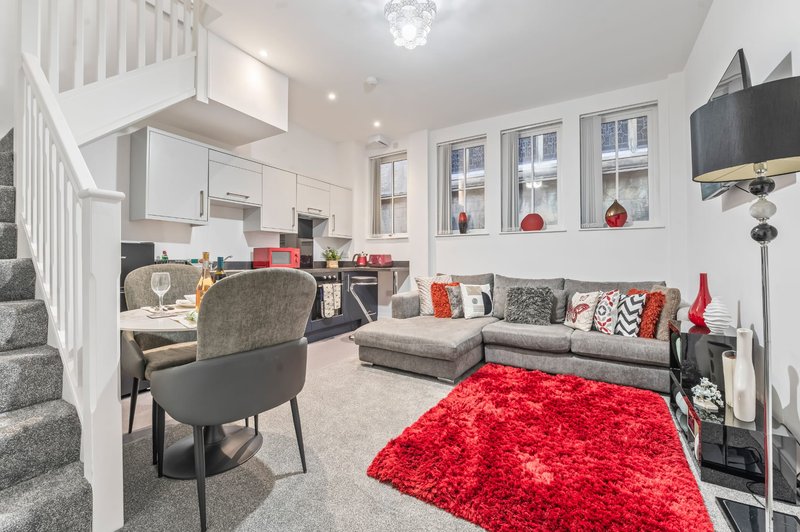 Open-plan living area with gray sofa, red rug, desk chair, and staircase to upper level