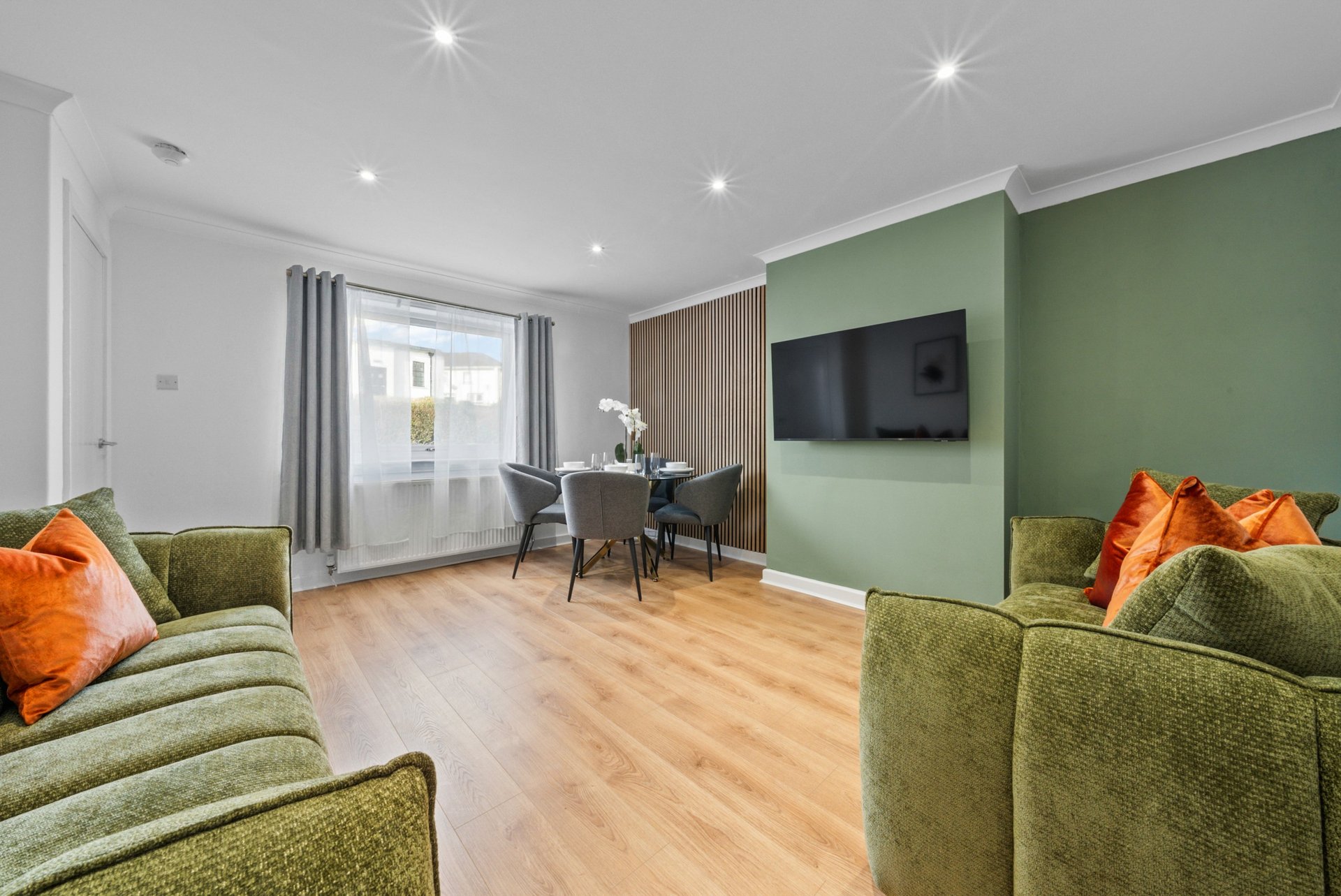Open-plan living room with green armchairs, orange cushions, wall-mounted TV, and wooden flooring