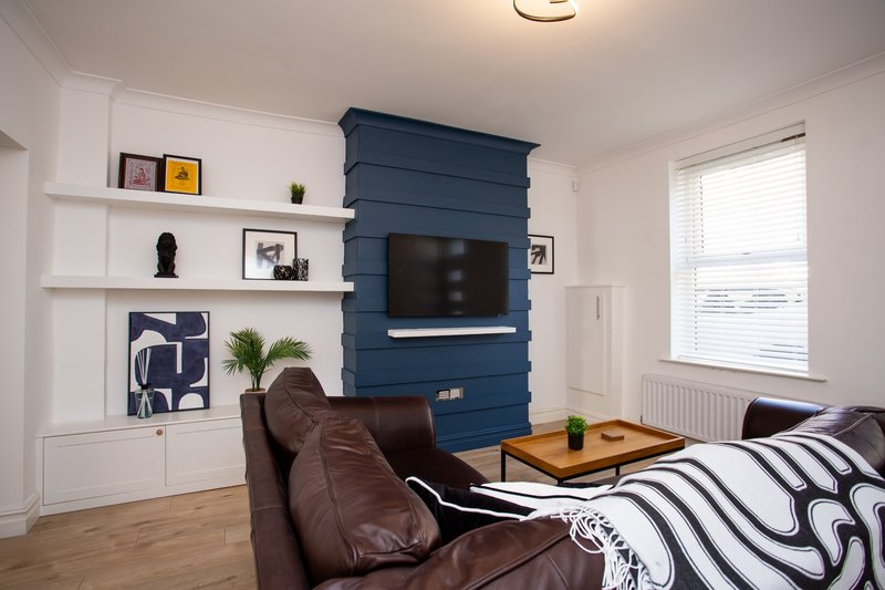 Living room with brown leather armchair, striped bedding, blue feature wall with mounted TV, white shelving and window.