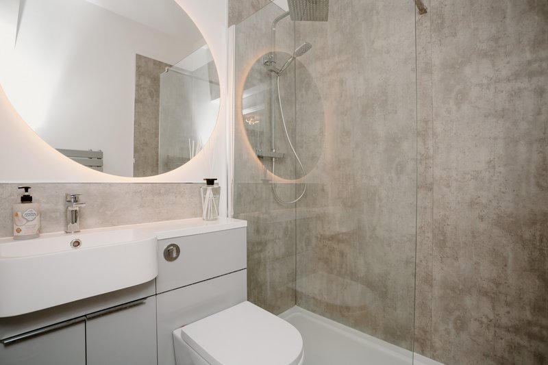 Modern bathroom with round illuminated mirror, white sink vanity, toilet, and walk-in shower with glass partition