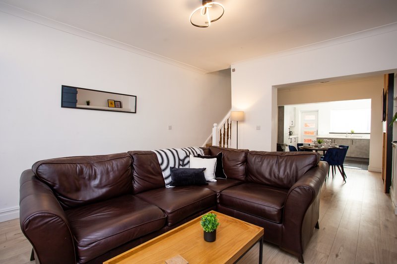 Modern living room with brown leather corner sofa, wooden coffee table, and open-plan kitchen visible through doorway