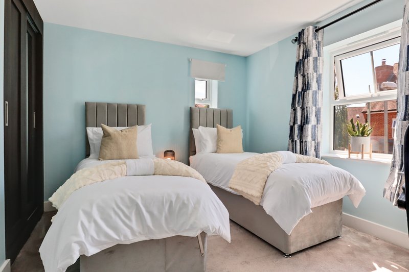 Bright twin bedroom with light blue walls, two single beds with white bedding and grey headboards, patterned curtains and window view