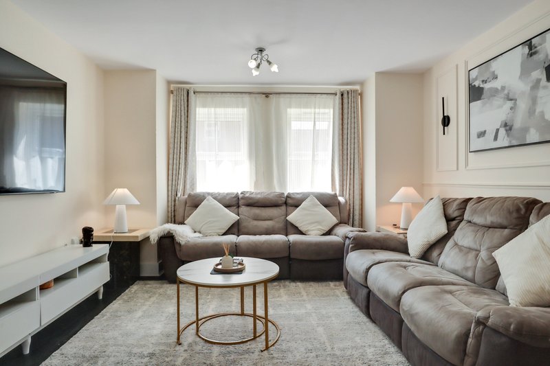 Modern living room with grey corner sofa, round coffee table, dual windows with curtains, and wall-mounted TV
