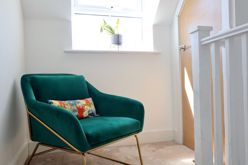 Bright corner room with teal upholstered armchair, colorful cushion, window with flowers, and white railing
