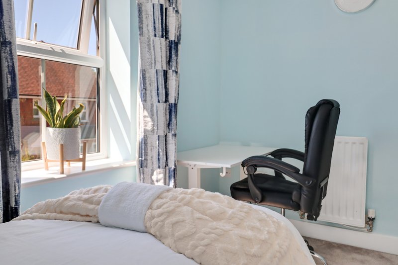 Bright bedroom with single bed, quilted beige throw, office chair, window with plant and patterned curtains