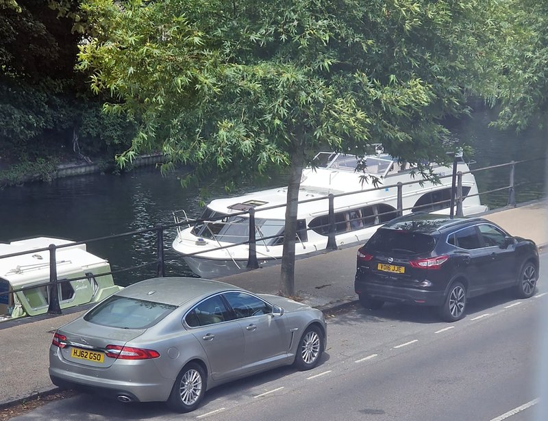Waterside property with moored boats, parked cars, mature trees and riverside walkway