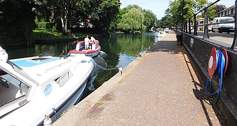 Waterside towpath with moored boats, trees, and residential buildings along canal