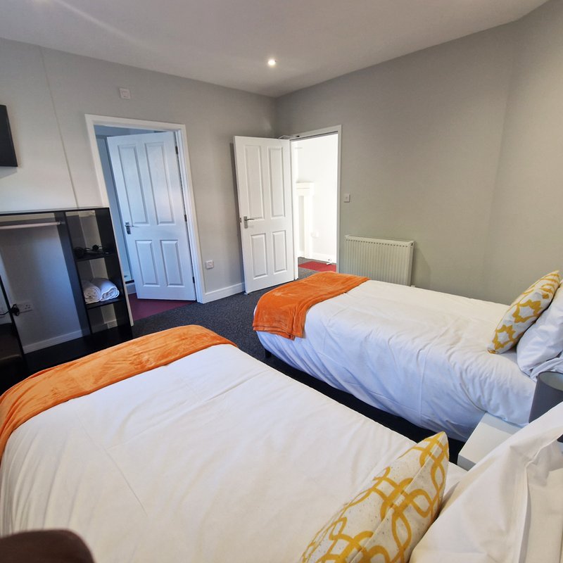 Twin bedroom with two single beds, orange throws, yellow patterned cushions, and doors to bathroom and hallway