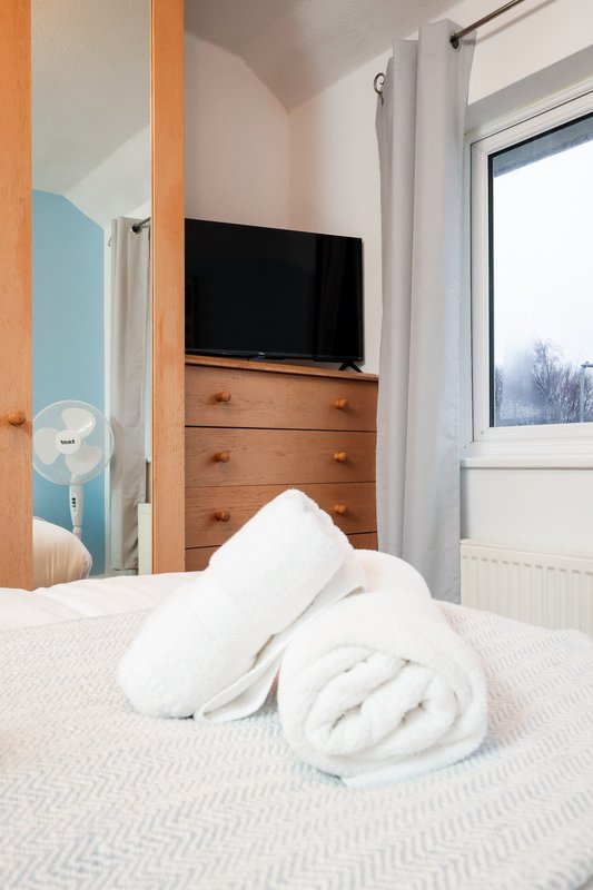 Bedroom with white bedding, wooden dresser, wall-mounted TV and bright window