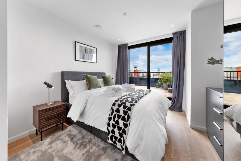 Modern bedroom with gray upholstered bed, wooden nightstand, and balcony view of cityscape