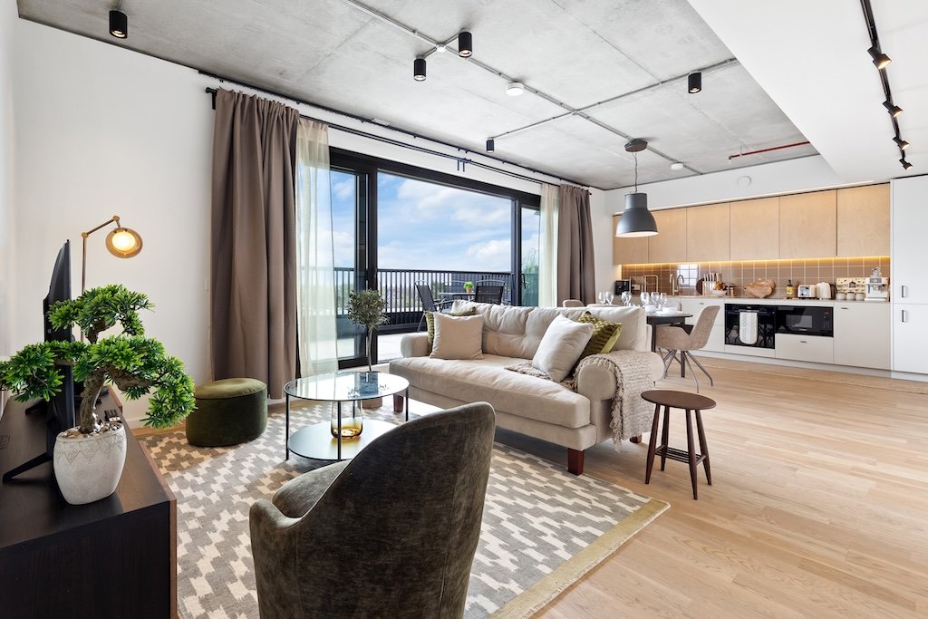 Modern open-plan living area with beige sofa, armchair, wooden flooring, and kitchen visible beyond