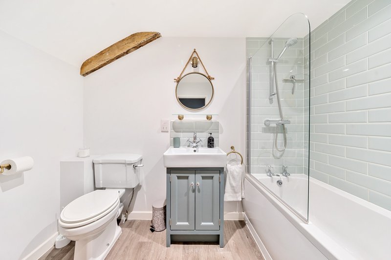 Modern bathroom with white subway tile, gray vanity, round mirror, toilet, and bathtub with glass enclosure