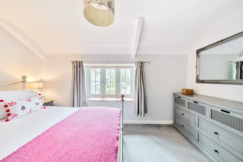 Bright bedroom with double bed, pink bedding, grey dresser, mirror and window with curtains
